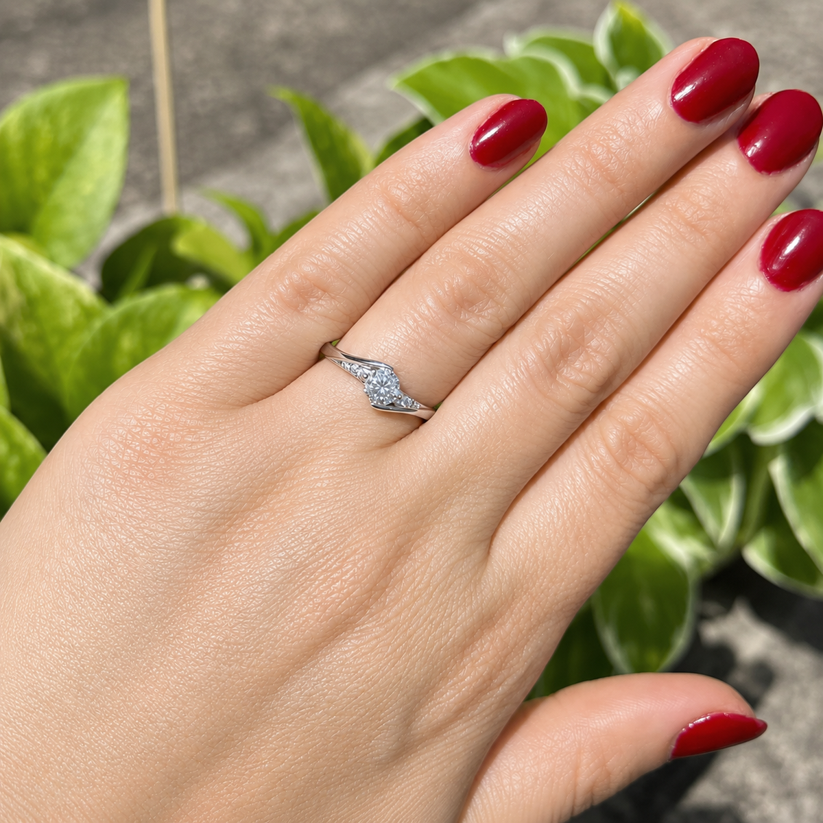 A silver ring with a marquise-cut diamond and smaller side stones, shown on a person's ring finger. The hand has dark red painted nails, and the background features blurred green houseplant leaves.