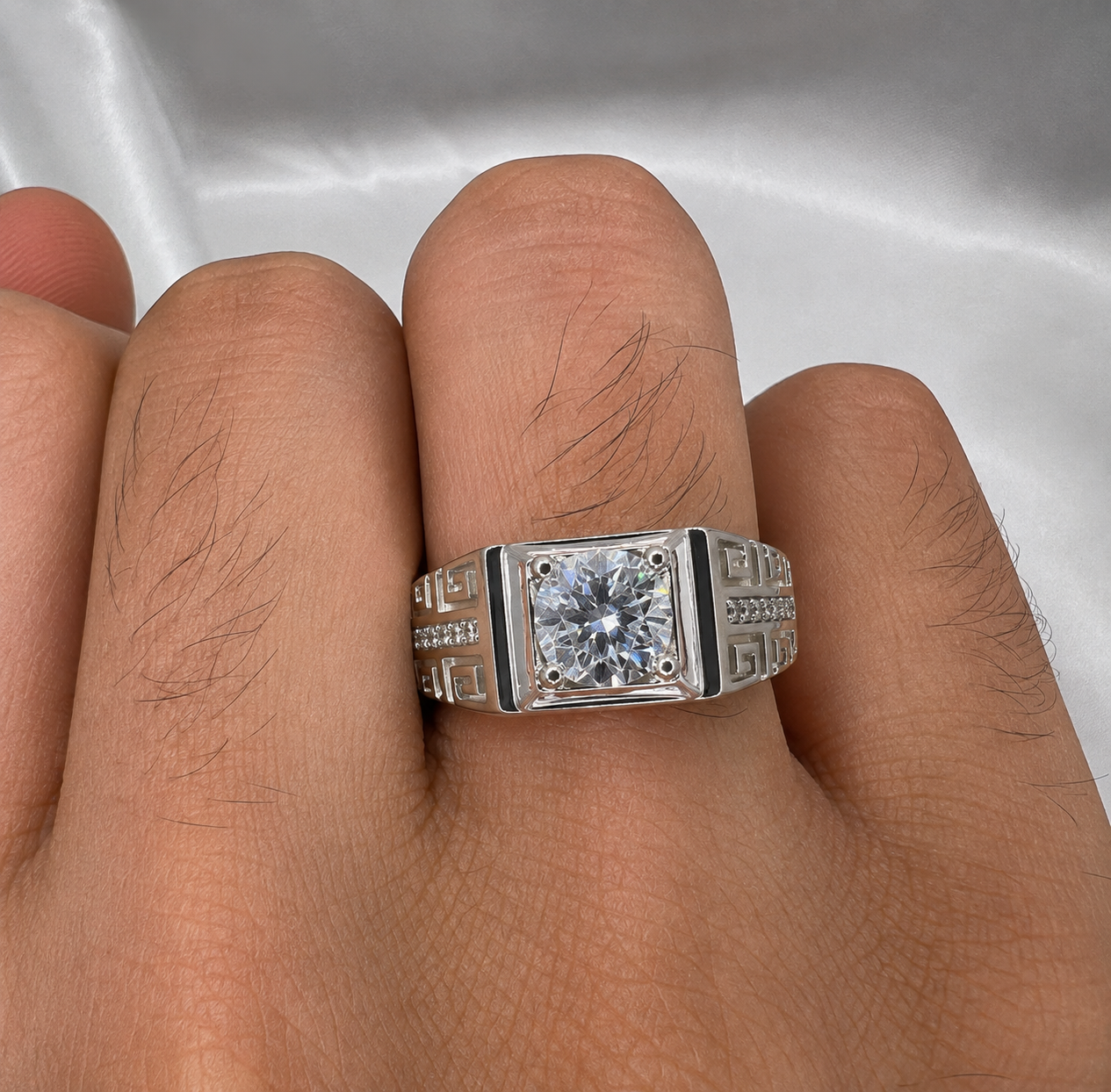 A silver-toned men's ring with a square-cut clear center stone and geometric side detailing, shown worn on a person's finger against a white satin background.