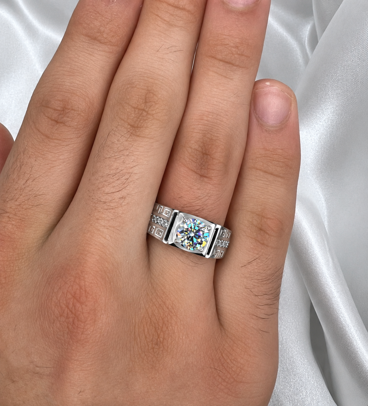 A close-up of a hand wearing a silver-toned ring with a large iridescent square gemstone and smaller stones along the band, set against a white satin fabric background.