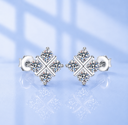 A pair of silver stud earrings with a geometric diamond-shaped design set with clear round gemstones, displayed on a light blue reflective surface.