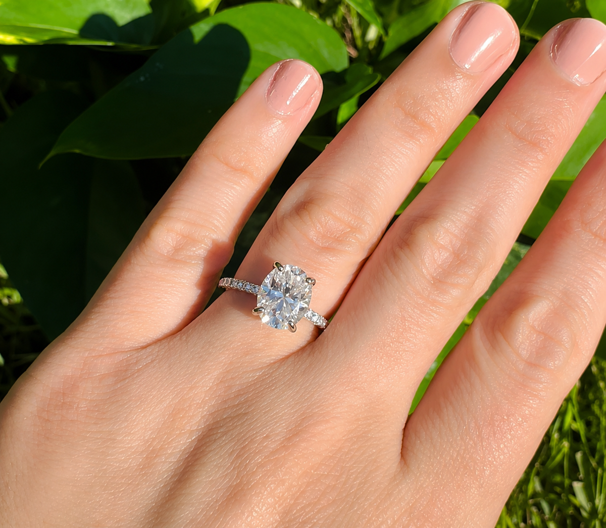 A close-up of a hand wearing a silver-toned ring with a large oval-cut clear center stone and small side stones, set against a background of green leaves.