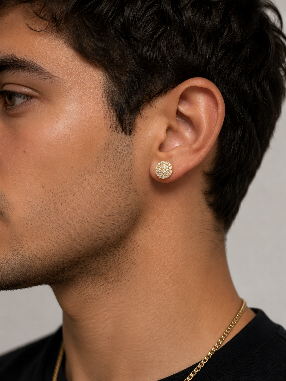 A close-up side profile of a man wearing a small, round, textured gold stud earring and a gold chain necklace against a neutral gray background.